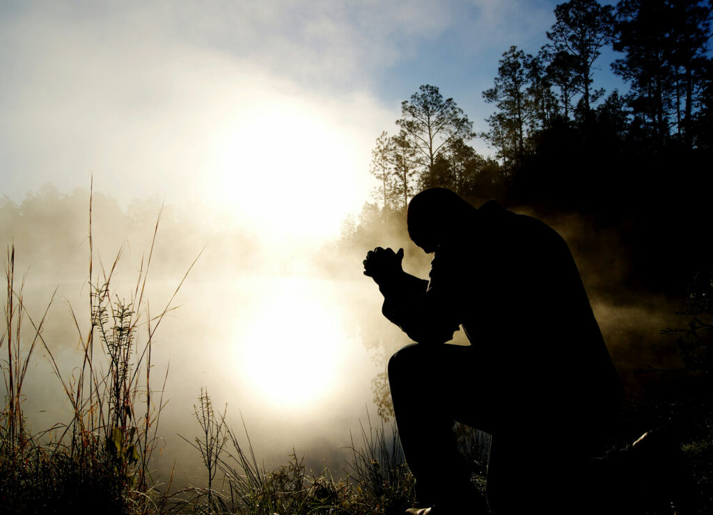 A person praying beside a quiet lake at sunrise